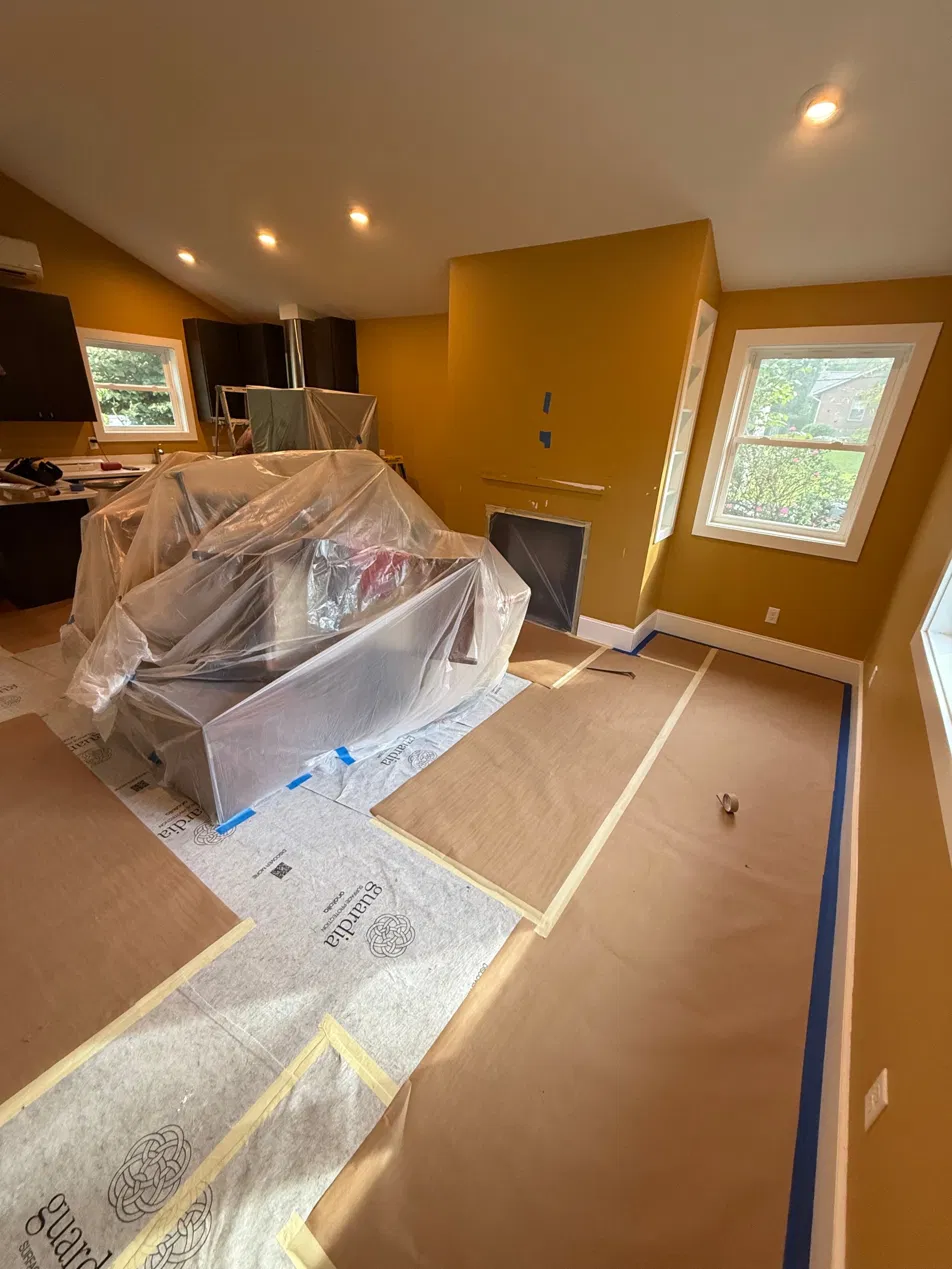 Living room furniture gathered and wrapped mid-room, floor protection taped to baseboards before painting