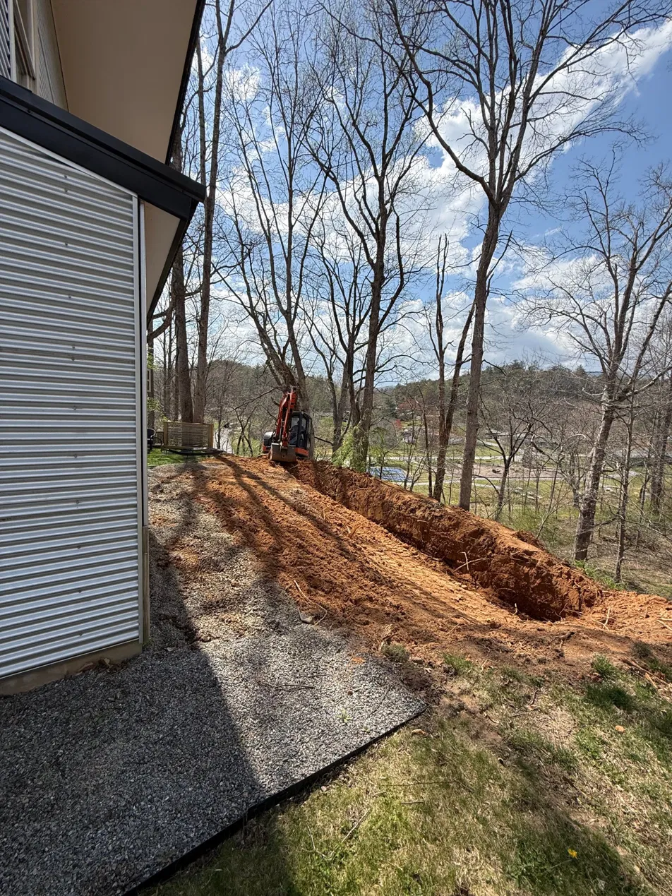 Footing trench opened alongside the existing house on the hillside, house metal siding visible at left